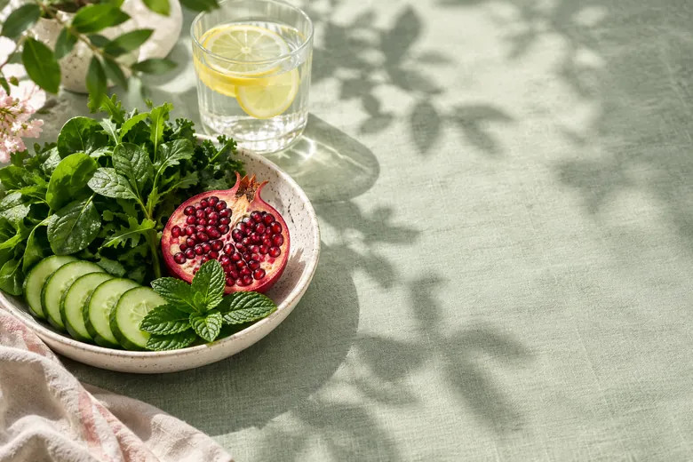 A bowl of leafy greens, cucumber, mint and a halved pomegranate beside a glass of lemon water on a linen cloth.